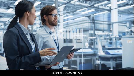 Portrait of an Experienced Male and Female Engineers Standing in Agency Room, Using Computer Devices and Discussing Production at a Modern Automated Electronics Manufacture with Robot Arms Stock Photo