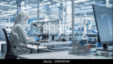 Modern Factory: Industrial Engineer Working on Desktop Computer in Clean Sterile Coveralls. Scientist Using a Microscope, Developing Advanced Solutions for High Tech Medical Vaccine and Gene Research Stock Photo