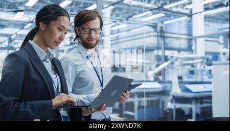 Portrait of Experienced Male and Female Engineers Standing in Agency Room, Using Computer Devices and Discussing Production at a Modern Automated Electronics Manufacture with Robot Arms Stock Photo