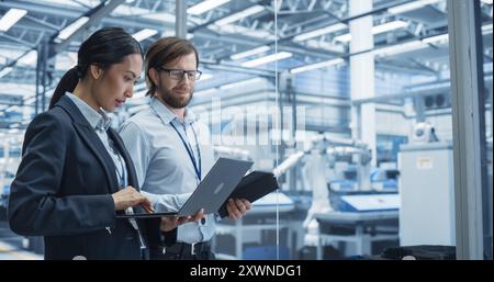Two Multiethnic Engineers Standing in Office, Using Laptop and Tablet Computers at an Electronics Factory. Machines are Undergoing Maintenance, Specialists Monitoring the Progress on Devices Stock Photo