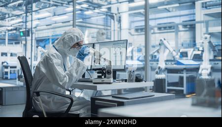 Modern Factory: Industrial Engineer Working on Desktop Computer in Clean Sterile Coveralls. Scientist Using a Microscope, Developing Advanced Solutions for High-Tech Medical Vaccine and Gene Research Stock Photo