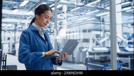 Portrait of Beautiful Electronics Manufacturing Technician Working on a Laptop Computer at a Factory. Asian Female Specialist Developing, Maintaining, Setting Up Programs for Machine Production Stock Photo
