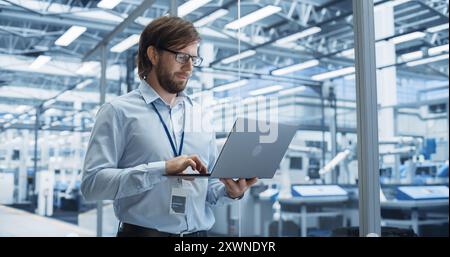 Portrait of a Handsome Engineer with Glasses Using Laptop Computer and Looking Around a Factory Facility with Equipment Producing Modern Electronic Components for Different Industries Stock Photo