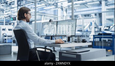 Software Developer Programming an Artificial Intelligence Neural Network on a Desktop Computer. Male Working in a Modern Technological Facility with an Automated Production Line with Robotic Arms Stock Photo