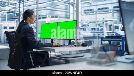 Female Robotics Technician Working on Desktop Computer with Green Screen Chromakey Mock Up Display in a Factory Office. Technological Research, Development Facility with Autonomous Assembly Line Stock Photo