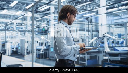 Experienced Male Engineer Standing in Office, Using Laptop Computer and Overlooking Production at Modern Automated Electronics Assembly Line with AI Support. Factory Executive at Work Stock Photo