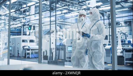 Development Factory Cleanroom: Engineers Wearing Coveralls and Goggles while Talking and Using Tablet Computer. Scientists Working in Advanced Electronics Manufacture with Precision Machinery Stock Photo