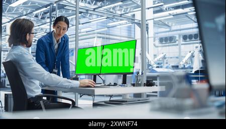 Portrait of a Young Male Engineer and Asian Team Leader Collaborating on a Project, Using Computer With Green Screen Chromakey On Display in a Factory with Equipment Producing Modern Technical Devices Stock Photo