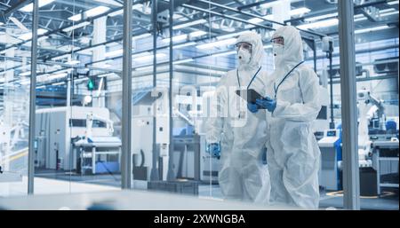 Development Factory Cleanroom: Engineers Wearing Coveralls and Goggles while Talking and Using a Tablet Computer. Scientists Working in Advanced Electronics Manufacture with Precision Machinery Stock Photo