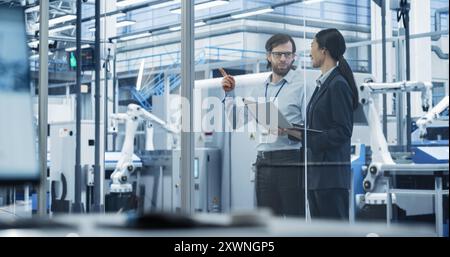 Portrait of a Young Engineer and Happy Asian Project Manager Using Laptop and Tablet Computers, Talking in a Factory Facility with Automated Robots Producing Modern Electronic Components Stock Photo