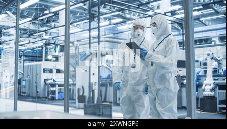 Development Factory Cleanroom: Engineers Wearing Coveralls and Goggles while Talking and Using Tablet. Scientists Working in Advanced Electronics Manufacture with Precision Machinery Stock Photo