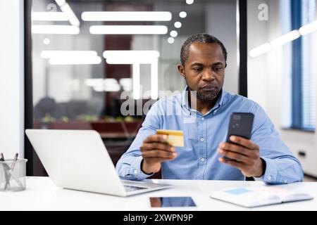 Serious businessman focused on completing online payment transaction using smartphone and credit card in office. Professional setting includes laptop, indicating work environment. Stock Photo