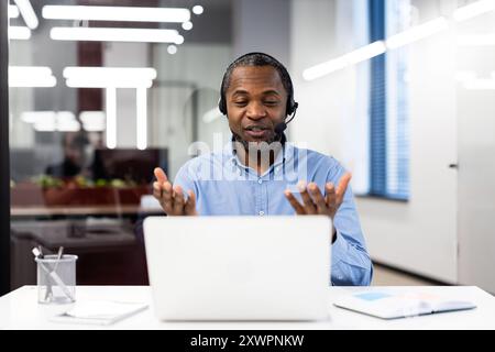 Businessman wearing headset having video call in modern office. He is smiling and gesturing while engaging in virtual meeting using laptop and phone technology Stock Photo