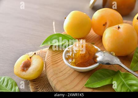 Jar with spoon filled with plum jam on wood cutting board and fruit around on wooden table. Elevated view. Stock Photo