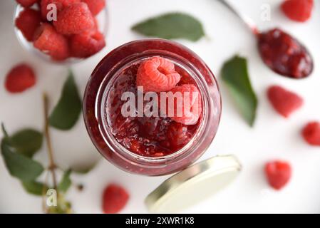 Detail of jar of raspberry jam on white table with fruit around it. Top view. Stock Photo
