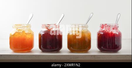 Collection of open jam jars of various with spoon on white wooden table and white isolated background. Front view. Stock Photo