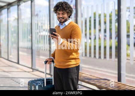 Confident man in orange sweater standing at bus stop, holding phone and suitcase. He is checking smartphone while waiting for public transport. Image conveys travel, communication, and relaxation. Stock Photo
