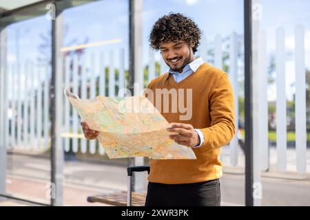 Happy young man with curly hair exploring city using map, standing at bus stop with suitcase. Captures travel, adventure, and navigation on bright sunny day. Stock Photo