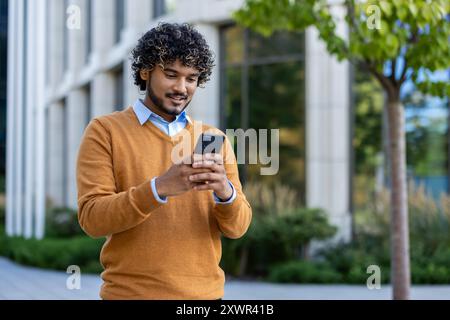 Young man enjoying outdoor work break, standing with phone in hand. Wearing casual sweater, he engages with technology in urban environment surrounded by nature, reflecting modern lifestyle Stock Photo