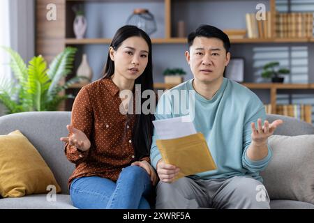 Asian couple sitting on sofa examining financial documents with concerned expressions. Couple appears to be discussing finances and paperwork in living room. Emotions convey worry and contemplation. Stock Photo