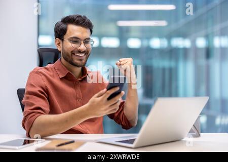 Young man in casual shirt expressing joy while using smartphone at office desk. Seated with laptop open, looking pleased and triumphant, indicating positive achievement. Atmosphere of success Stock Photo