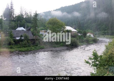 Picturesque village nestled by a gently flowing river surrounded by lush greenery and misty hills in the Karpaten region. The scene exudes tranquility and represents a serene and peaceful rural life. Stock Photo