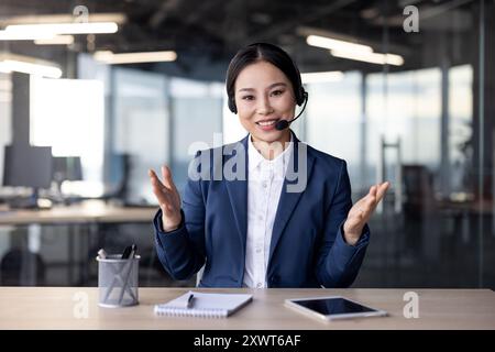 Asian businesswoman wearing headset smiling, talking on video call in modern office, connected with phone and tablet. Stock Photo