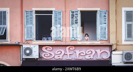 Kid looking out of a window on a colorful house front above "Safran" restaurant in Nice, France Stock Photo