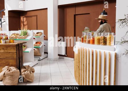 In sustainable local supermarket is a male shopkeeper standing at checkout counter. Salesman wearing a hat is arranging items over at cashier desk in bio store. Fresh, eco-friendly produce shown. Stock Photo