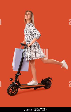 Young woman with shopping bags riding electric scooter on orange background Stock Photo