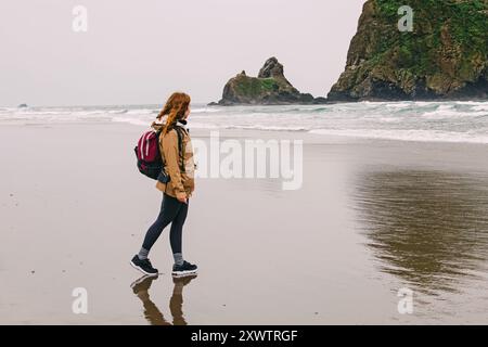 Woman walking along a sandy beach with a backpack near rocky cliffs on a cloudy day Stock Photo