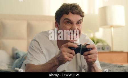 Playful Caucasian European young man lying on bed playing console holding joystick video games fun weekend home enjoy guy hobby leisure hands Stock Photo
