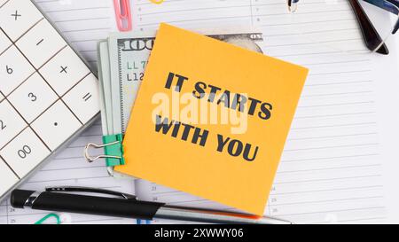 New Year Resolution News Concept. IT STARTS WITH YOU phrase on a yellow sticker on American banknotes on the background of a business notebook Stock Photo