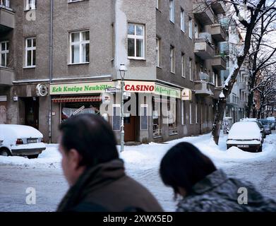 A wintery street view in Berlin-Neukölln featuring the Eck Kneipe Neukölln corner pub and a casino. The snowy sidewalks and two pedestrians create a typical urban winter atmosphere in this German neighborhood. Stock Photo