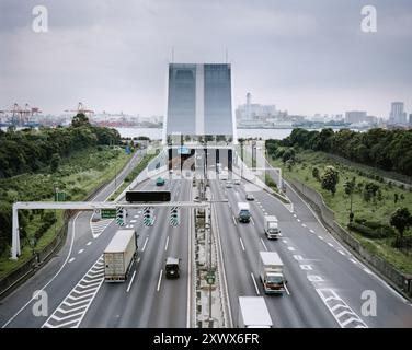 Vehicles moving on a highway as it enters a tunnel in Odaiba, Tokyo, Japan. The image captures the urban transportation infrastructure and greenery surrounding the area, highlighting the busy city life. Stock Photo