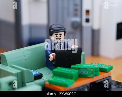 Tambov, Russian Federation - August 18, 2024 A Lego businessman minifigure sitting on a couch and working on a laptop behind a table with money on it. Stock Photo