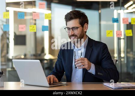Confident business professional using laptop in modern office setting. Engaged in work while holding drink, conveying focus and relaxation. Stock Photo