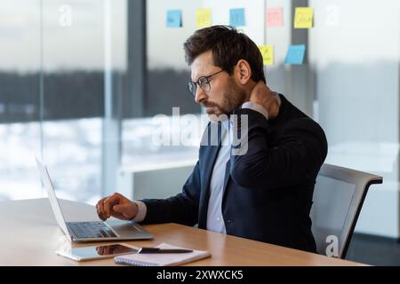 Businessman in suit sitting at desk using laptop, appearing uncomfortable due to neck pain. Office with focused work atmosphere illustrates concept of work-related stress and physical discomfort Stock Photo