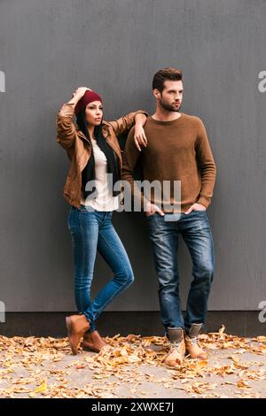 Enjoying autumn style. Beautiful young couple bonding to each other and looking away while leaning at the grey wall with fallen leaves laying around t Stock Photo