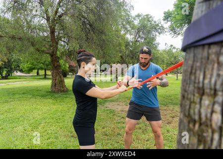 Two individuals performing a strength training exercise using resistance bands in a lush green park, engaging in an outdoor fitness routine focused on Stock Photo