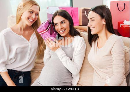 What is inside? Beautiful young pregnant woman holding gift box near ear and smiling while two friends sitting close to her on the couch Stock Photo