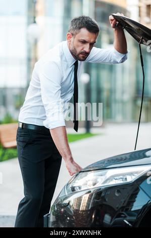 Having some car troubles. Serious young businessman looking inside of the vehicle hood while standing near his car Stock Photo
