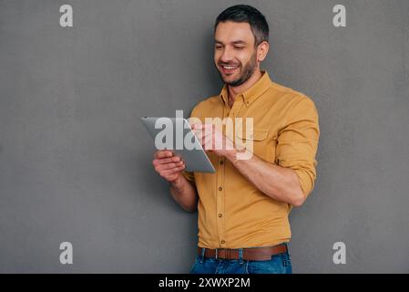 Examining his brand new tab. Handsome mature man holding digital tablet and looking at it with smile while standing against grey background Stock Photo
