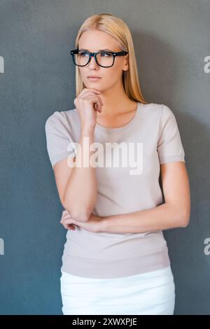 Thoughtful beauty. Thoughtful young blond hair woman holding hand on chin and looking away while standing against grey background Stock Photo