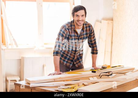 Smiling woodworker. Cheerful young male carpenter leaning at the wooden table with diverse working tools laying on it Stock Photo
