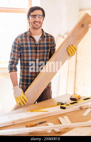 Dreams are born at my workshop. Confident young male carpenter holding wooden plank and smiling while standing in his workshop Stock Photo