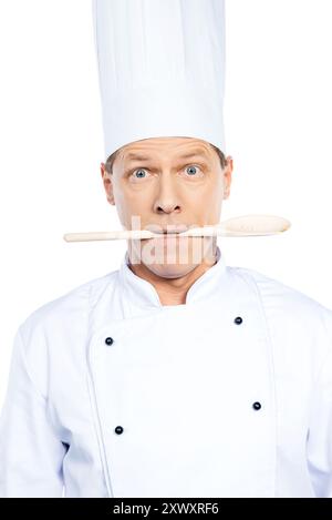 What to cook? Surprised mature chef in white uniform holding wooden spoon in his mouth while standing against white background Stock Photo