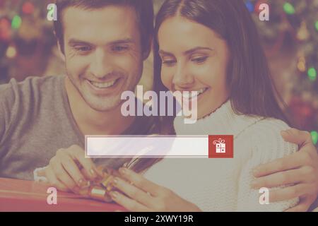 Presents search. Happy young woman opening a gift box while her boyfriend sitting close to her and smiling with Christmas decoration in the background Stock Photo