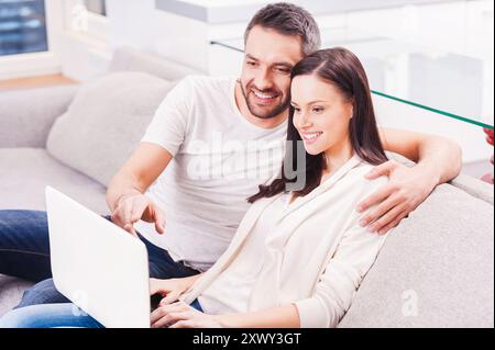 Surfing the net together. Beautiful young loving couple bonding to each other and looking at the laptop with smile while sitting on the couch together Stock Photo