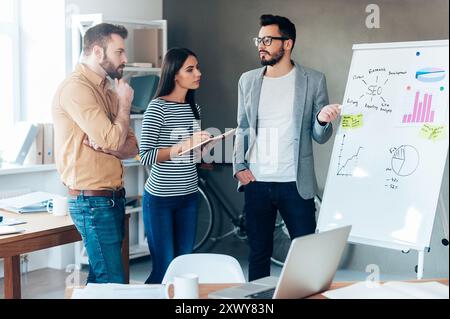 Discussing some new project together. Confident young man standing near whiteboard and pointing it with smile while his colleagues standing near him Stock Photo